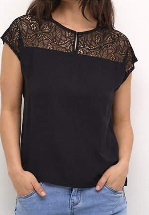 Femme portant un haut noir sans manches avec un empiècement en dentelle délicate et des manches courtes, associée à un jean en denim bleu clair, les mains dans les poches.