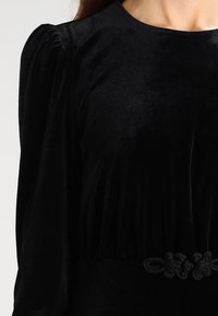 Vestido de terciopelo negro con una textura suave, con mangas fruncidas, escote redondo y un cinturón decorativo anudado en la cintura.