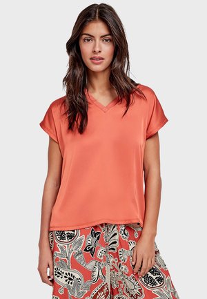 Kobieta o długich ciemnych włosach nosi jednolity koralowy top z krótkim rękawem i dekoltem w serek oraz koralową spódnicę z wzorem w czarno-białe kwiaty i ptaki.