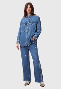 Camicia di denim con decorazioni in borchie argentate, dotata di due tasche frontali e una vestibilità rilassata. Abbinata a pantaloni di denim coordinati adornati con borchie.