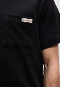 Μαύρο κοντομάνικο πουκάμισο με τσέπη στο στήθος που φέρει λευκή ετικέτα με το λογότυπο Calvin Klein, φορεμένο από ένα άτομο με εμφανές γυμνό μπράτσο.