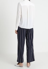 Witte blouse met lange mouwen en een achterkant met schouderpas, gecombineerd met marineblauwe wijde broek met gestreepte patronen in roze en witte verticale lijnen. Schoenen zijn instappers.