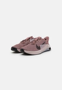Calzado deportivo Nike en malla rosa claro con acentos negros, suelas de goma planas y cierre con cordones; presenta un diseño distintivo.