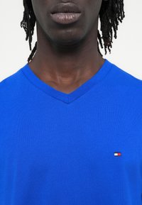 Μπλε μπλουζάκι με v λαιμόκοψη από μαλακό ύφασμα, με μικρό οριζόντιο λογότυπο σε κόκκινο, λευκό και ναυτικό στο αριστερό μέρος του στήθους. Υφή στην επιφάνεια.