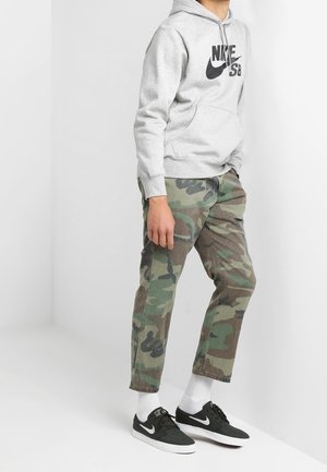 Personne portant un sweat à capuche Nike SB gris, un pantalon camouflage, des chaussettes blanches et des baskets Nike noires, debout contre un mur blanc.