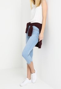 Jean cropped bleu clair avec un léger effilochage à l'ourlet, associé à un top sans manches blanc et un sweat à capuche bordeaux noué. Des baskets blanches complètent le look.