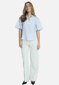 Blouse bleu clair à manches courtes et col, avec une finition texturée, associée à un pantalon large blanc et des chaussures noires à bout pointu.