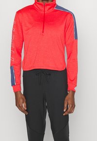 Röd kvartdragspullover med marinblå axelaccent, mjuk textur, långa ärmar och logodetalj på vänster arm. Tillsammans med svarta byxor.