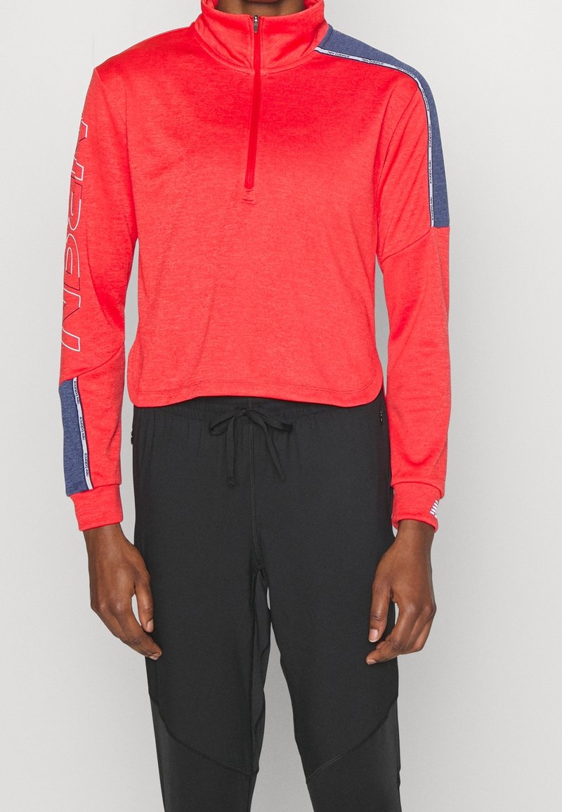 Röd kvartdragspullover med marinblå axelaccent, mjuk textur, långa ärmar och logodetalj på vänster arm. Tillsammans med svarta byxor.