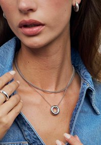 Silverfärgad kedjehalsband med en cirkulär medaljong som har en strukturerad yta. Modellen bär en jeansskjorta. Ring synlig på höger hand.