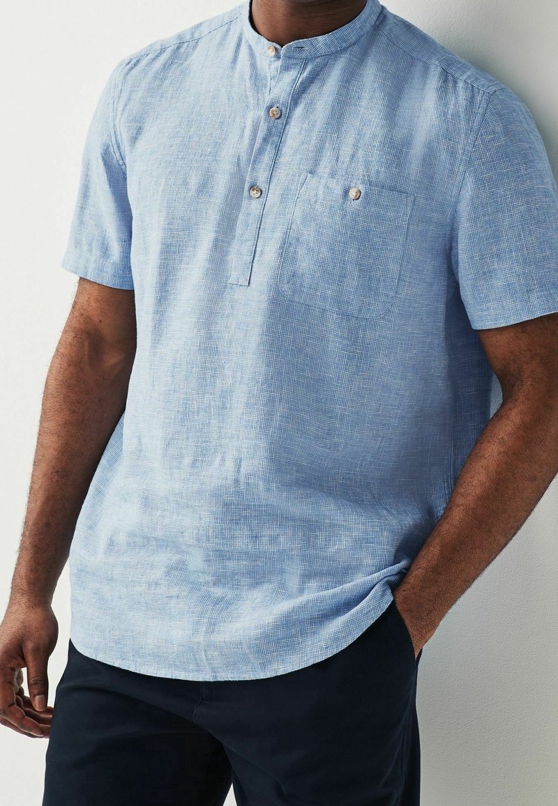 Homme portant une chemise bleu clair à manches courtes avec col mandarin, trois boutons et une poche poitrine, associée à un pantalon foncé.
