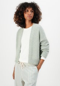 Lichtgroene gebreide cardigan met een relaxte fit, open voorkant en geribde mouwen, gedragen over een wit shirt en gecombineerd met gestreepte broeken.