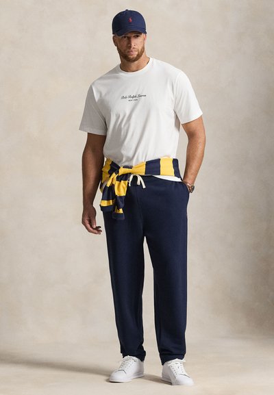 Άνδρας που φοράει λευκό T-shirt Polo Ralph Lauren, σκούρο μπλε φόρμα, λευκά αθλητικά παπούτσια, σκούρο μπλε καπέλο και πουλόβερ με κίτρινες και σκούρο μπλε ρίγες δεμένο γύρω από τη μέση.