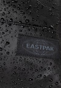 Black waterproof fabric with a textured surface, featuring numerous water droplets. A black Eastpak logo is stitched onto the material.