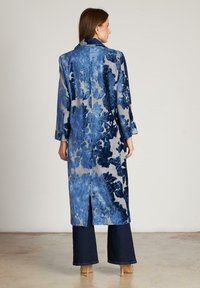 Abrigo floral largo con un patrón en azul y gris. Cuenta con un collar, mangas largas y una abertura en la parte trasera. Combinado con pantalones anchos oscuros y tacones.