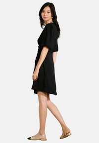 Robe noire à manches ballons, longueur genou, en tissu léger. Associée à des chaussures à talons slingback beiges et noires.