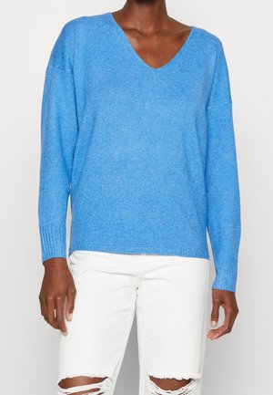 Dunkelhäutige Person in hellblauem V-Ausschnitt-Pullover und weißen, zerrissenen Jeans vor schlichter Hintergrund.
