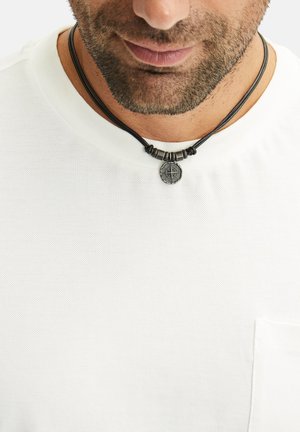 Zwarte leren ketting met een ronde metalen hanger met een kompasontwerp, geplaatst tegen een gestructureerd wit T-shirt.