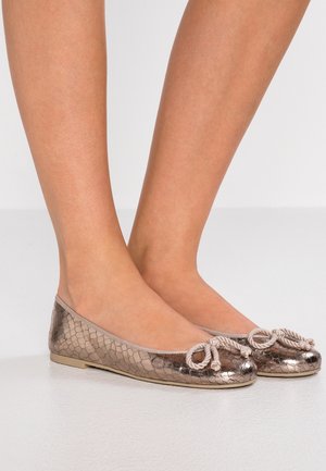 Stopy w metalicznych, brązowych balerinach o fakturowanej powierzchni, z ozdobnymi beżowymi kokardami na wierzchu, na jasnym tle.