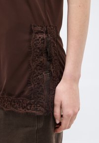 Δεξί χέρι δίπλα σε σκούρο καφέ σατέν ένδυμα με διαφανές δαντελένιο τελείωμα πάνω από σκουρόχρωμο παντελόνι, σε ανοιχτόχρωμο χέρι μπροστά από ανοιχτό φόντο.