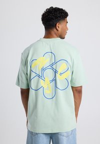 Light green cotton t-shirt featuring a large floral design in blue outline with yellow accents on the back, casual fit and short sleeves.