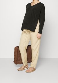 Svart, oversized, långärmad stickad tröja med V-ringning; beige, löst sittande byxor med knytband i midjan; vävd brun väska; platta rep-sandaler.