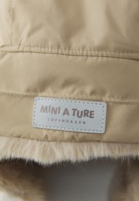 Dettaglio di una giacca puffer beige con un morbido rivestimento in pelliccia all'orlo e un'etichetta grigio chiaro con la scritta "MINI A TURE COPENHAGEN" impresso in grigio scuro.
