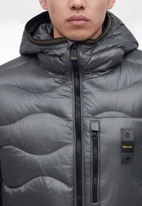 Gros plan d'une personne portant une veste matelassée grise avec capuche, fermeture éclair frontale, poche poitrine verticale et étiquette Blauer sur la poitrine.