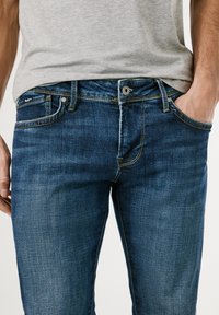 Donkerblauwe denim jeans met een gladde textuur, voorzien van gele stiksels, een vijf-pocket ontwerp en een metalen knoopsluiting bij de taille.