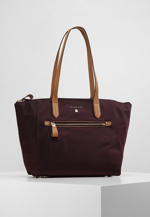 Ciemnofioletowa torba typu tote z jasnobrązowymi skórzanymi uchwytami i kieszenią na zamek, logo Michael Kors nad kieszenią, prezentowana na białej powierzchni.