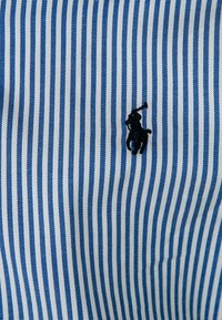 Modrobílé pruhované látka s texturovaným povrchem. Obsahuje malý černý emblém s koněm a jezdcem.