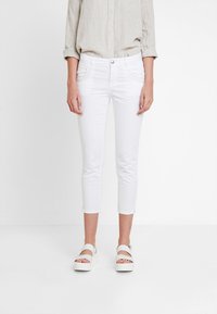 Mujer vestida con una camisa beige de manga larga, pantalones ajustados recortados blancos y sandalias de plataforma blancas, contra un fondo blanco.