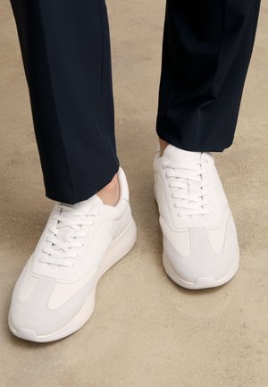 Personne portant des baskets blanches à lacets et un pantalon bleu marine, debout sur un sol beige texturé.