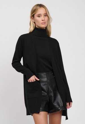 Blonde Frau trägt schwarzen Rollkragenpullover, lange schwarze Strickjacke mit Taschen und schwarze Leder-Shorts, posiert vor einfachem hellem Hintergrund.
