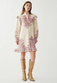 Cremefarvet kjole med intrikate pink og blå blomster mønstre, flæser på ærmerne og krave med knapper, afsluttet med en flæset kant.