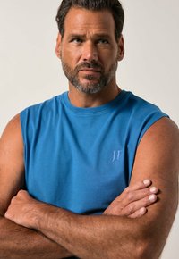 Man med kort mörkt hår och skägg iklädd en blå ärmlös skjorta, med armarna över kors och tittande lätt åt sidan mot en neutral bakgrund.