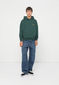 Felpa verde in cotone con tasca frontale e logo giallo, abbinata a jeans a gamba larga blu chiaro e sneakers nere con dettagli bianchi.