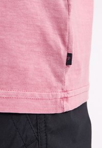 Rosa Baumwoll-T-Shirt mit abgerundetem Saum und einem kleinen schwarzen Stoffetikett an der Seite. Die Textur wirkt glatt mit einem leichten Stretch.