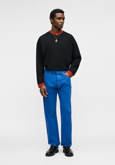 Άνδρας όρθιος με το χέρι στην τσέπη, φοράει μαύρο πουκάμισο με μακριά μανίκια και κόκκινους μανσέτες, φωτεινό μπλε παντελόνι, μαύρα παπούτσια και κολιέ.