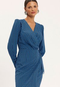 Niebieska sukienka o kroju kopertowym z wzorem w diamenty, długimi rękawami i wiązaniem w talii. Gładki materiał o lekkim połysku. Srebrne kolczyki w kształcie kół.
