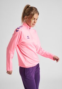Maglietta sportiva rosa con mezza zip e maniche lunghe, con logo del marchio viola e design a chevron sulle spalle. Tessuto liscio e leggero.