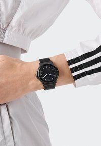 Mustad mustri kell ümmarguse näoga, minimalistlike tunni tähistega, tekstuuriga kummirihma ja roostevabast terasest korpusega.