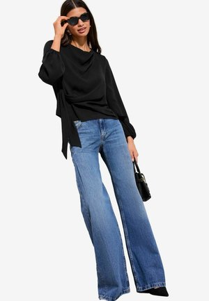 Frau trägt eine schwarze Langarmbluse, weit geschnittene blaue Jeans, schwarze Sonnenbrille und trägt eine kleine schwarze Handtasche.