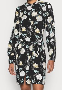 Černé šaty s dlouhým rukávem a knoflíkovým zapínáním, s žlutým a bílým květinovým potiskem, s opaskem na zavázání v pase a s rozparkem vpředu, které nosí osoba.