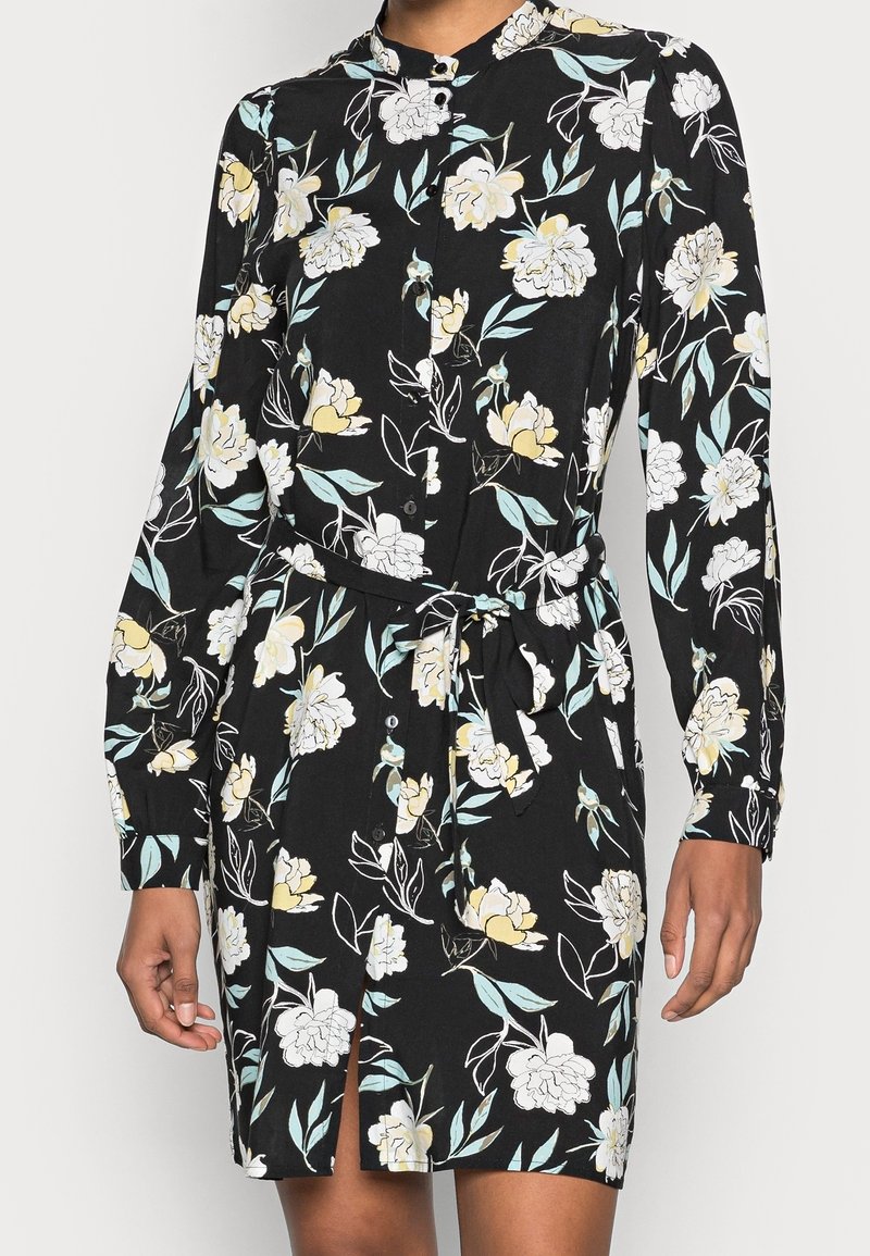 Černé šaty s dlouhým rukávem a knoflíkovým zapínáním, s žlutým a bílým květinovým potiskem, s opaskem na zavázání v pase a s rozparkem vpředu, které nosí osoba.