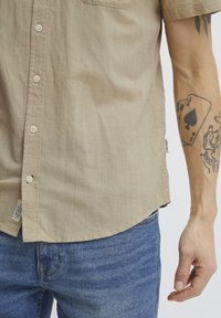 Camisa de botones de manga corta en un beige claro, con un bolsillo, una tela texturizada y un dobladillo curvado, combinada con jeans de mezclilla azul.
