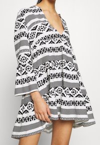 Robe à motifs noir et blanc avec un col en V, des manches longues évasées et une jupe évasée présentant des motifs géométriques et des rayures contrastées.