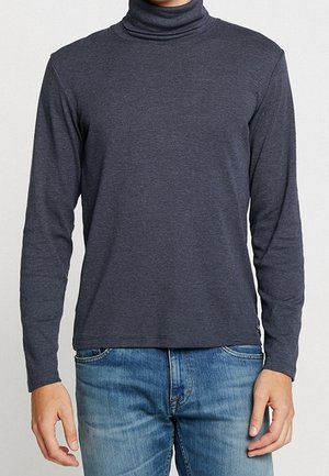 Mężczyzna ubrany w ciemnoszary golf z długim rękawem i jasnoniebieskie dżinsy, stojący z ramionami swobodnie opuszczonymi wzdłuż ciała.