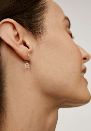 Pendiente de aro plateado con un colgante ovalado, que presenta un detallado patrón de pequeñas piedras, cuelga de una oreja perforada. Tono de piel visible.