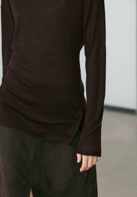 Brązowa bluzka z długim rękawem o dopasowanym kroju i guzikach po bokach. Tkanina wydaje się miękka, o gładkiej fakturze, na tle ciemniejszej spódnicy.